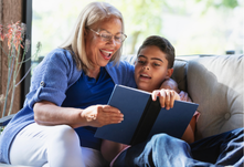 An older adult wearing glasses reads with a younger person on a couch